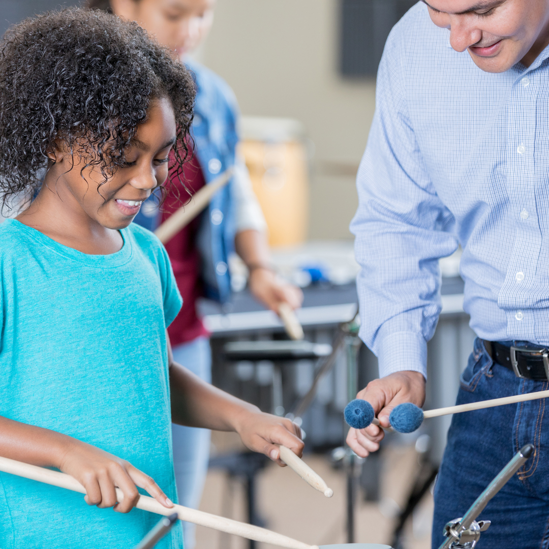A child learning to play the drums is shown what to do by their teacher