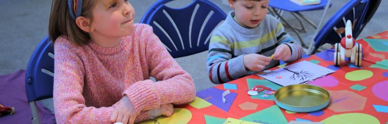 Picture of a little girl, and boy at a craft table, cutting out shapes and gluing them to paper